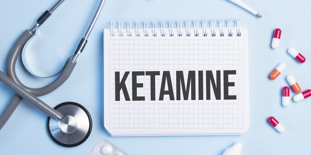 The word ketamine written on a white notepad on a blue background near a stethoscope, syringe, electronic thermometer and pills. Medical concept